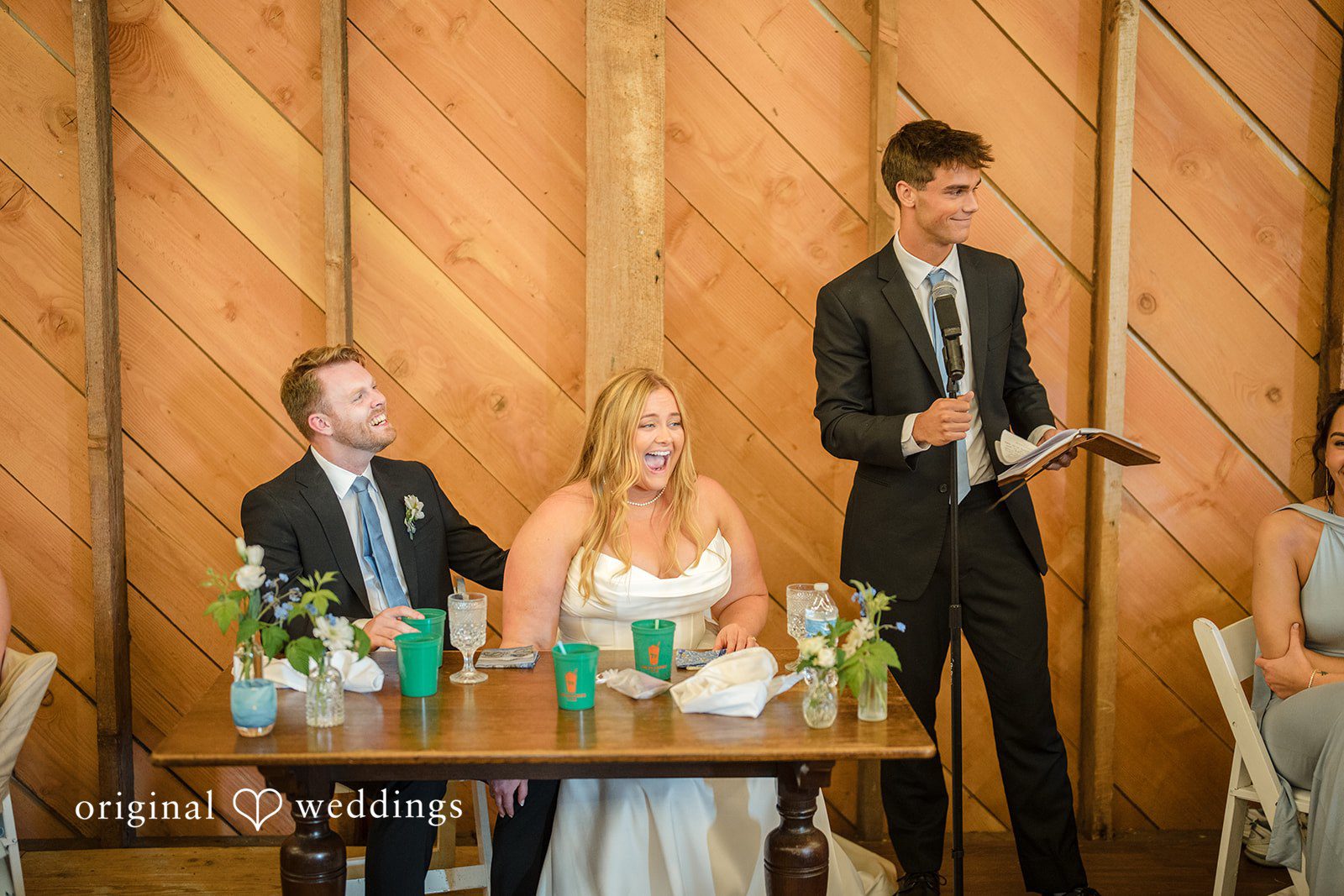 Madelyn + Connor A man giving a speech at a wooden podium while a bride and groom listen from a decorated table.