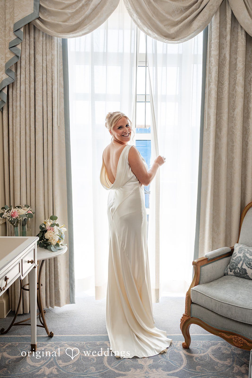 Maggie + Taylor A bride wearing a sleek white wedding gown poses gracefully next to a window.