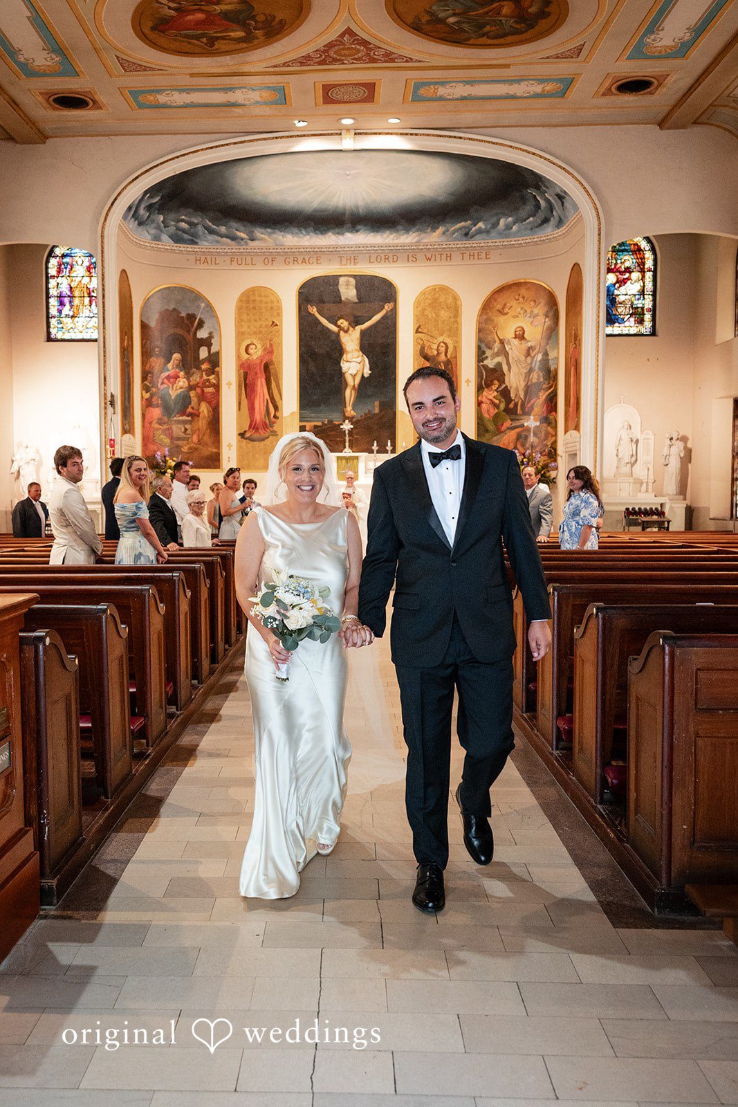 Maggie + Taylor A newlywed couple smiles as they walk down the church aisle together.