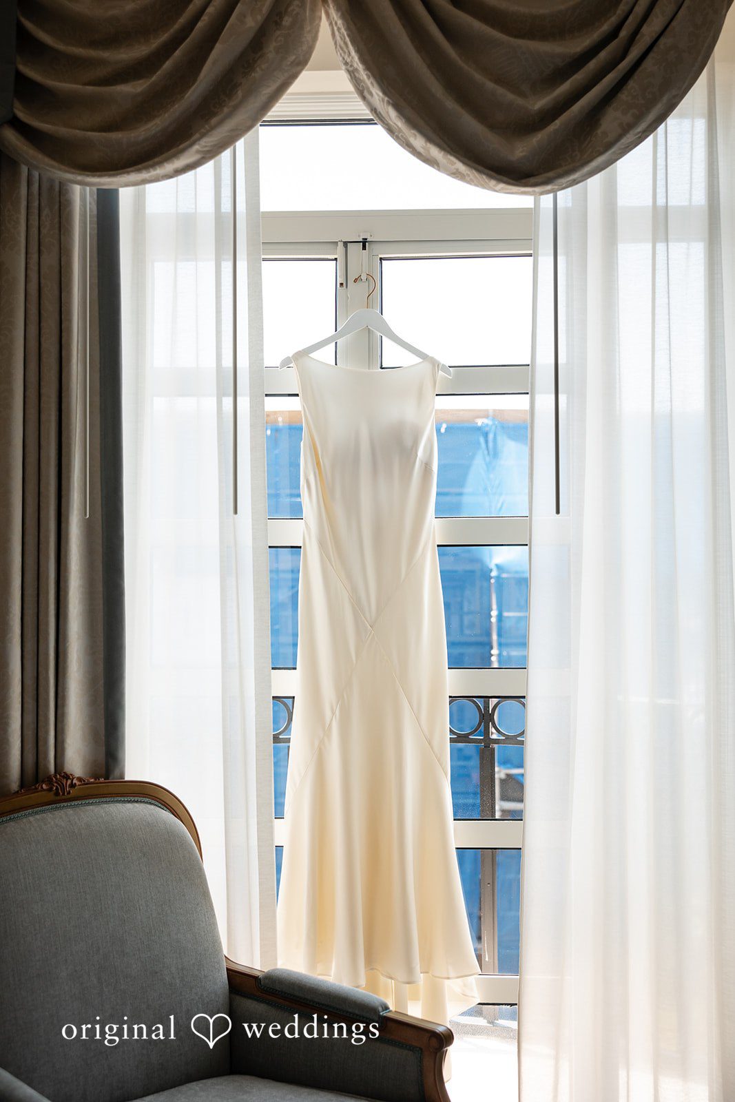 Maggie + Taylor A white wedding dress hangs elegantly in front of a window.
