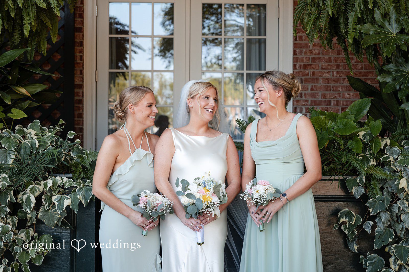 Maggie + Taylor A bride poses smiling with two bridesmaids in sage green gowns.