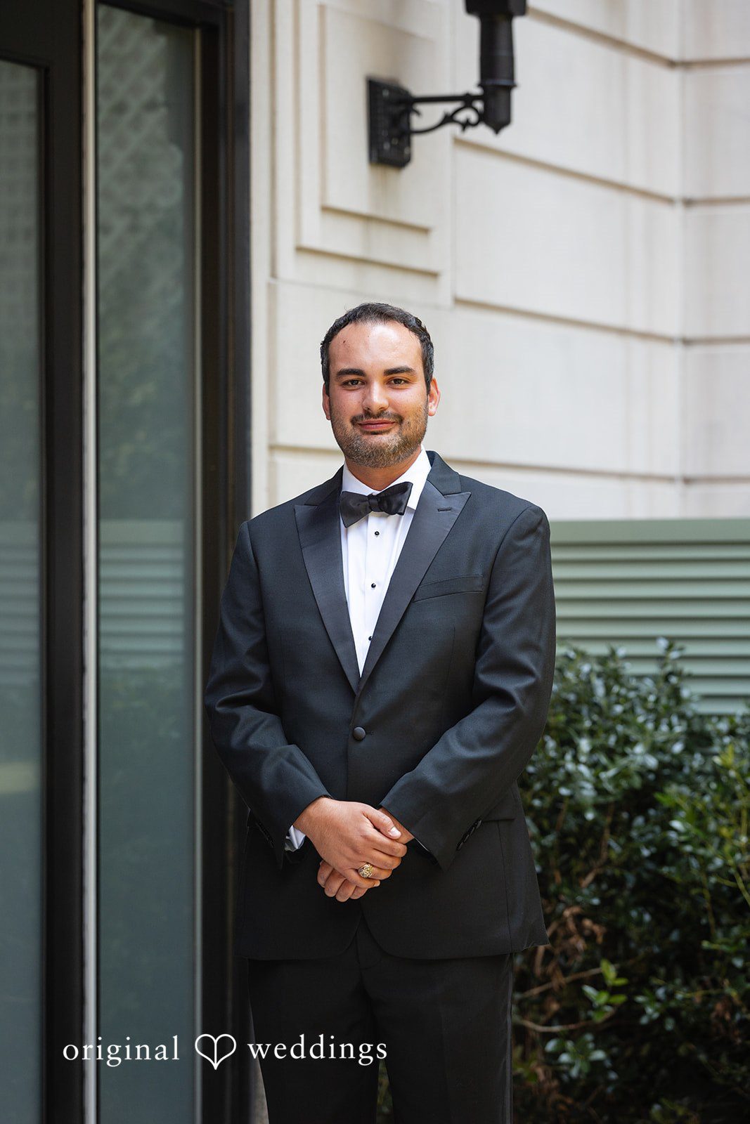 Maggie + Taylor A groom wearing a black tuxedo smiles while standing in front of a building