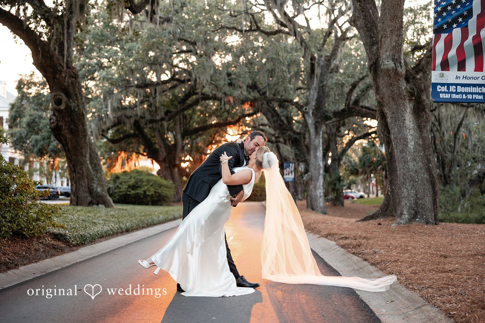 Maggie + Taylor A couple shares a kiss on a road framed by large trees.
