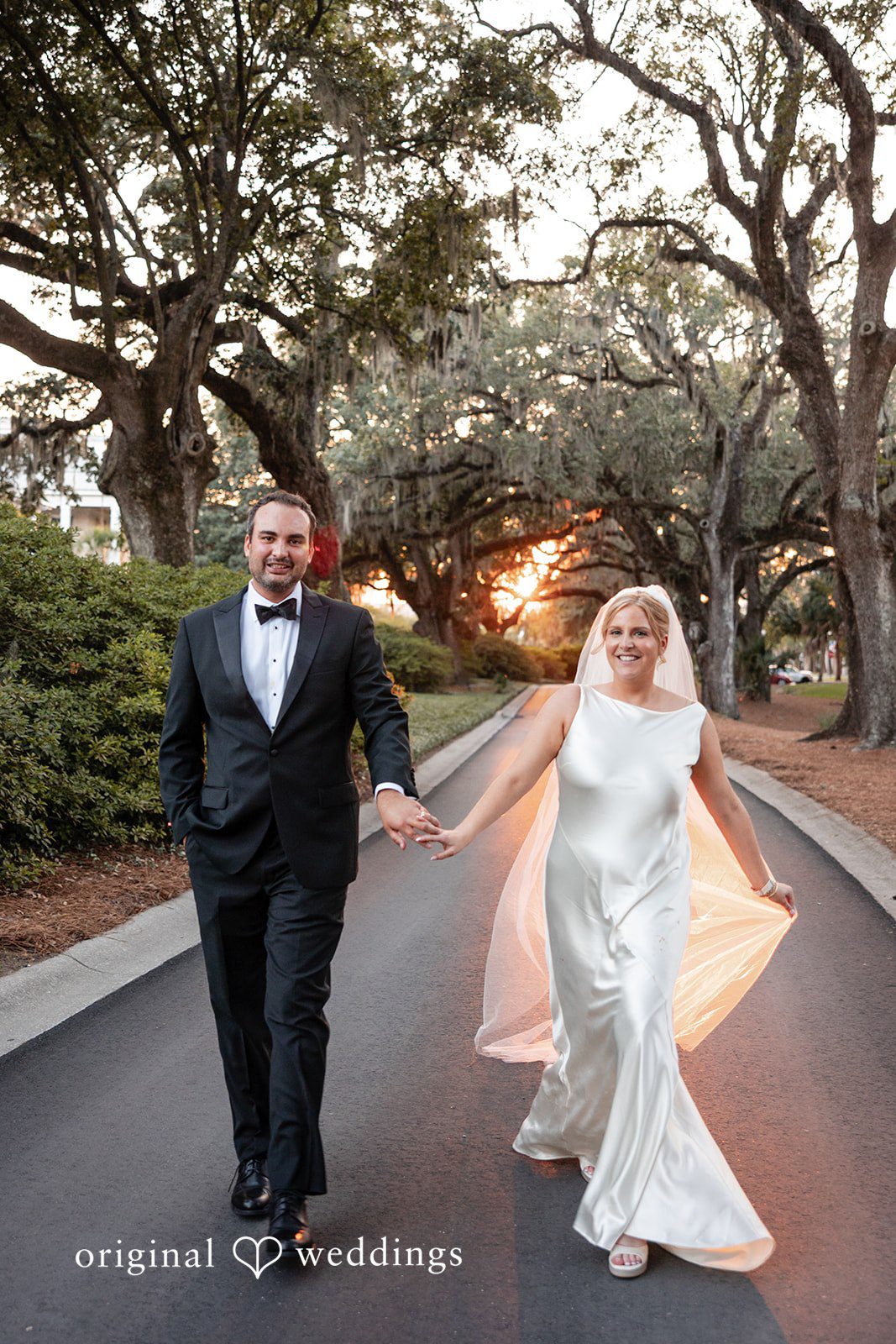 Maggie + Taylor A bride and groom joyfully walk hand-in-hand down a paved road.