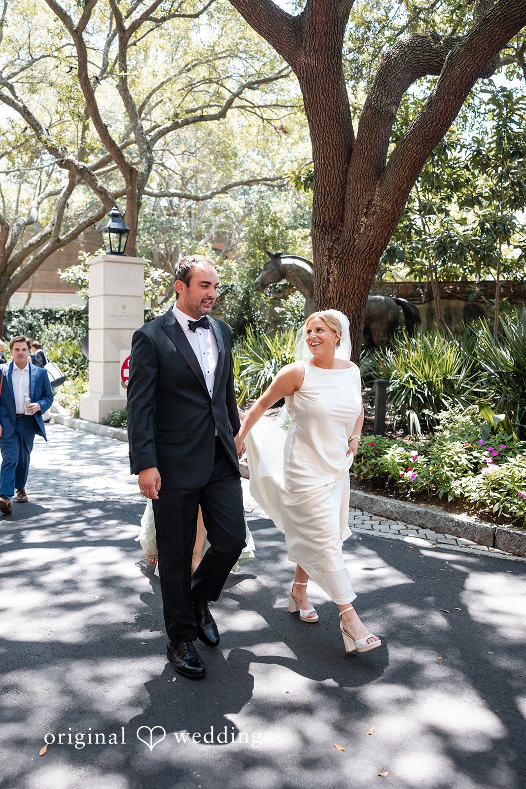 Maggie + Taylor A bride and groom hold hands while walking along a sunlit pathway.