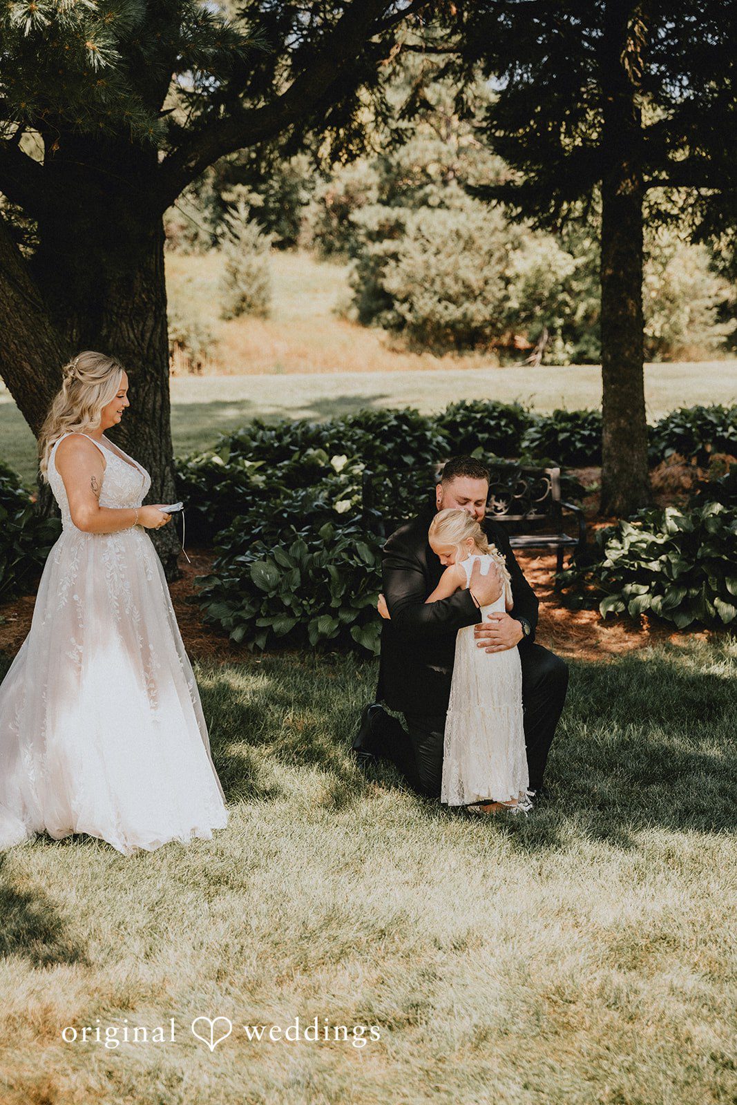 Samantha + Peyton Bride standing while the groom sits with little child on the grass beside her.