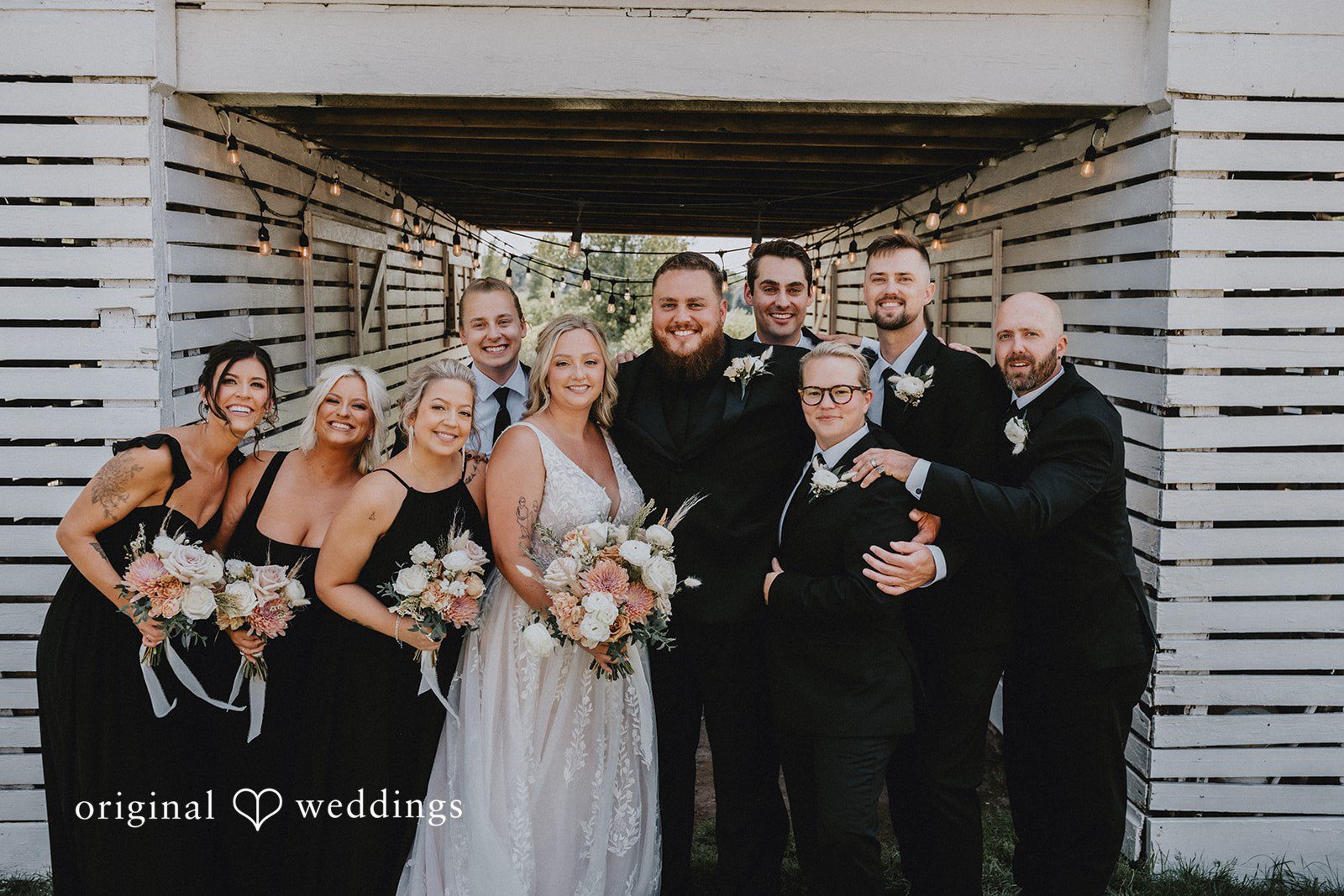 Samantha + Peyton Wedding party smiling and posing together beside a white wooden structure.
