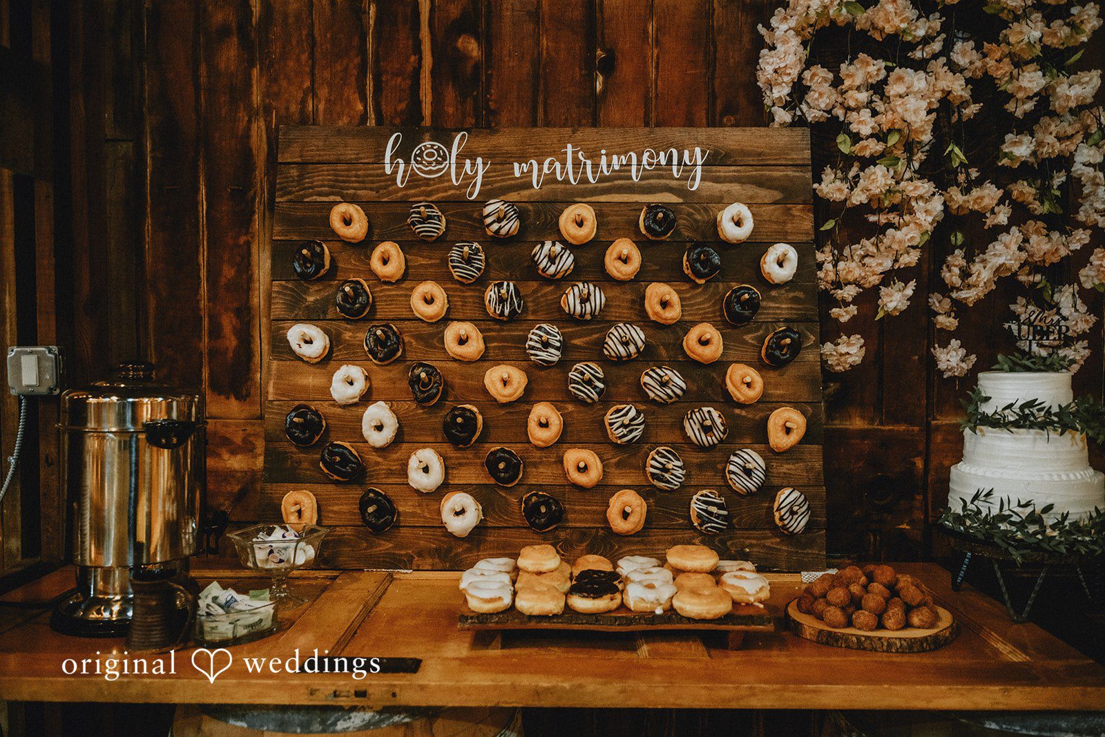 Samantha + Peyton Rustic wooden wall display featuring rows of assorted donuts below a "holy matrimony" sign next to a wedding cake.