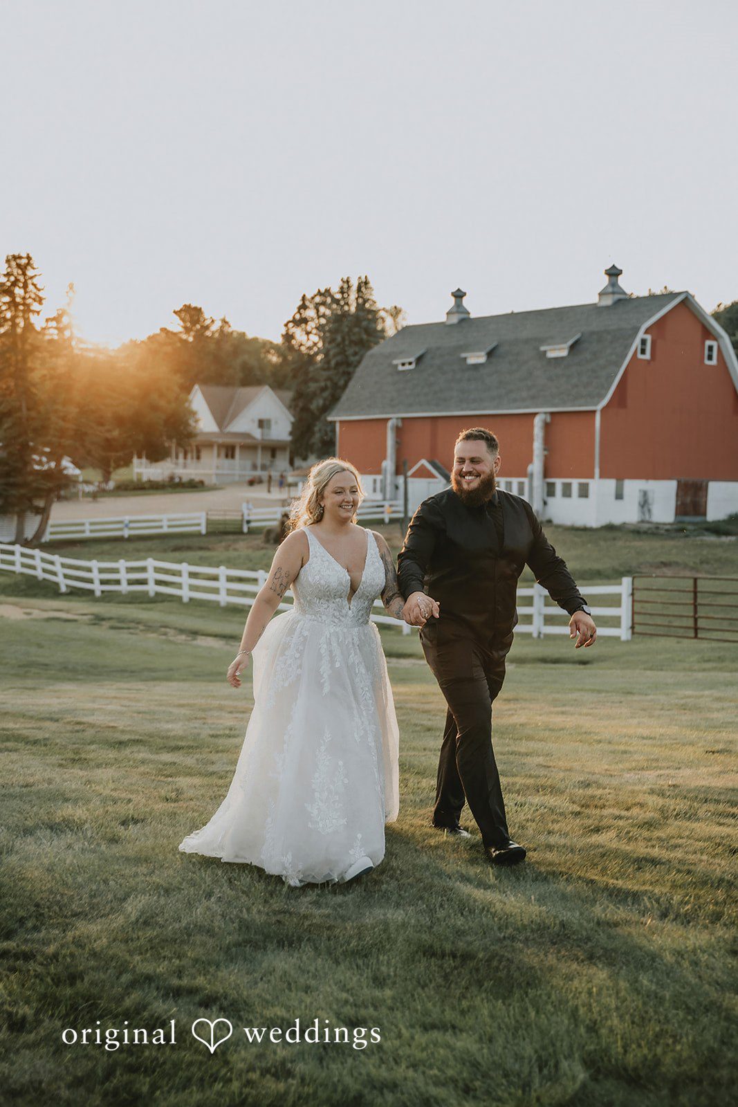 Samantha + Peyton Newlyweds running hand in hand across a field toward a red barn.