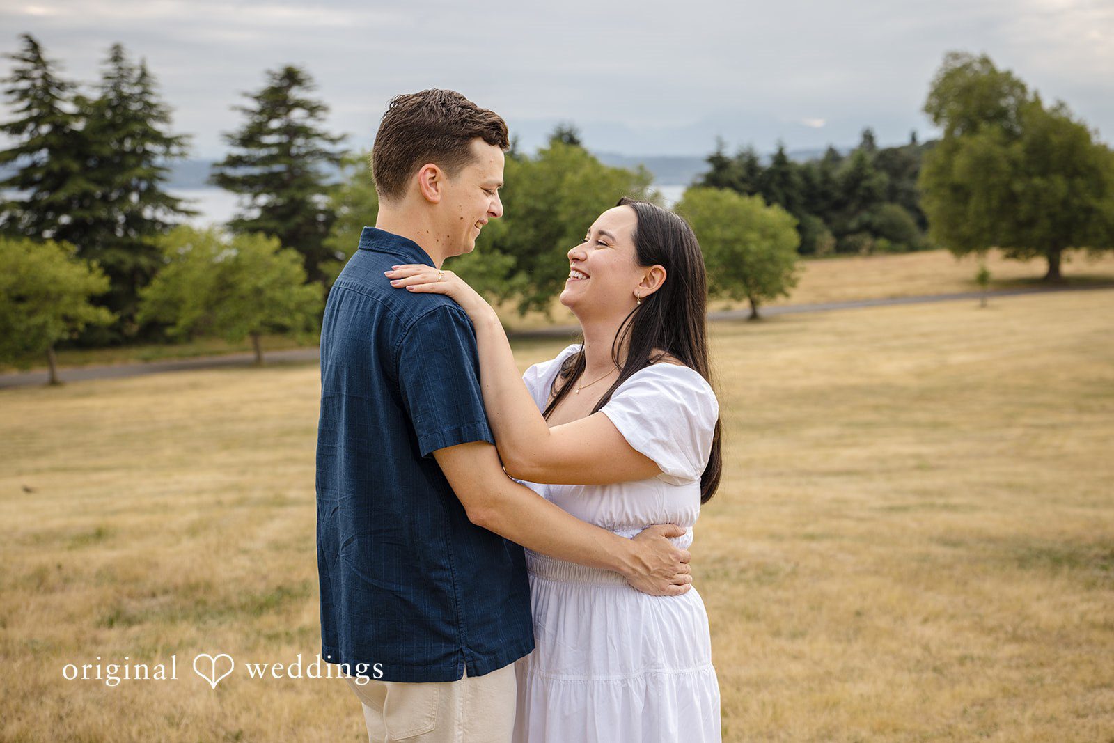 A man and woman smiling and looking at each other while embracing in a grassy field.