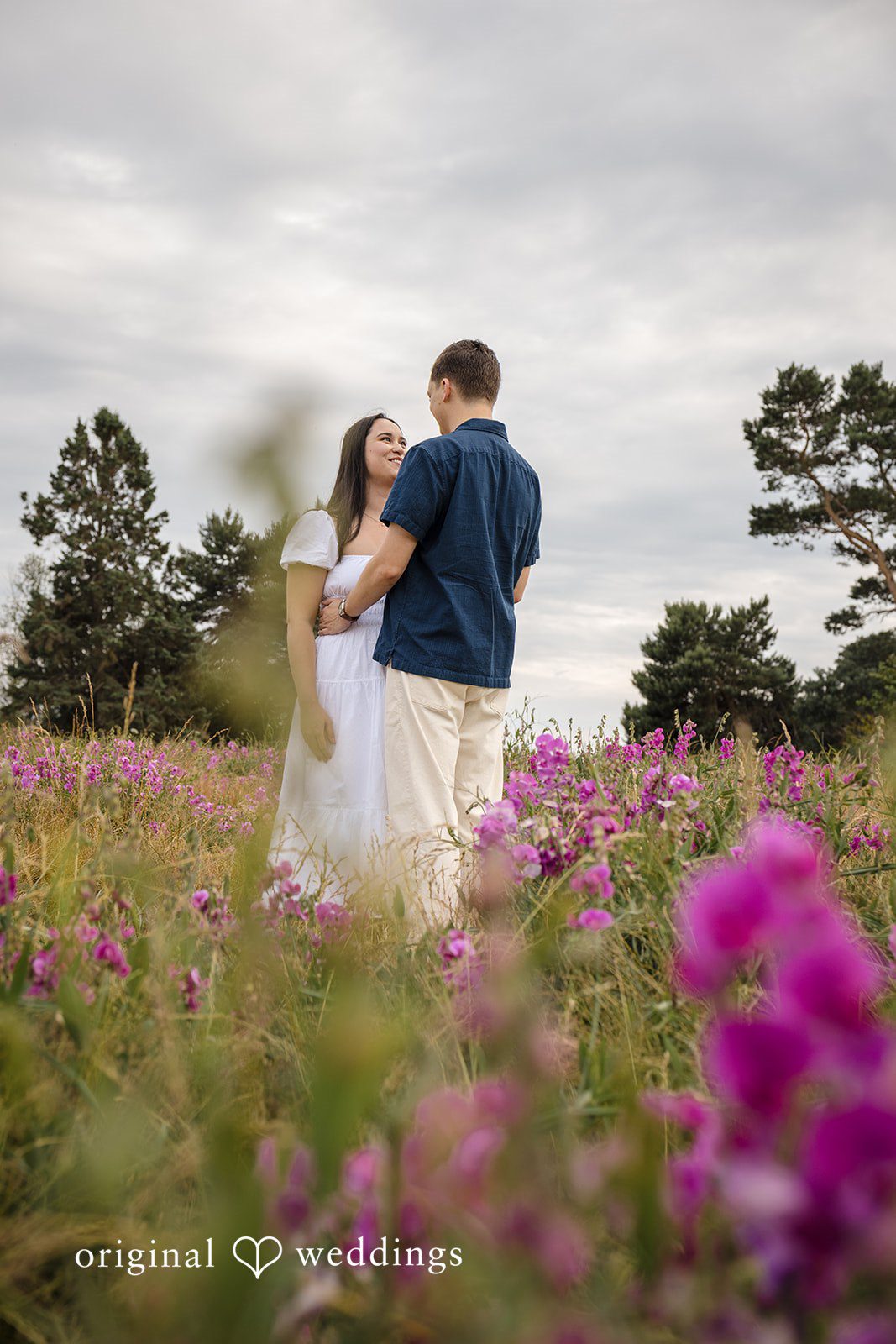 A couple standing together on a hill surrounded by tall pink wildflowers.