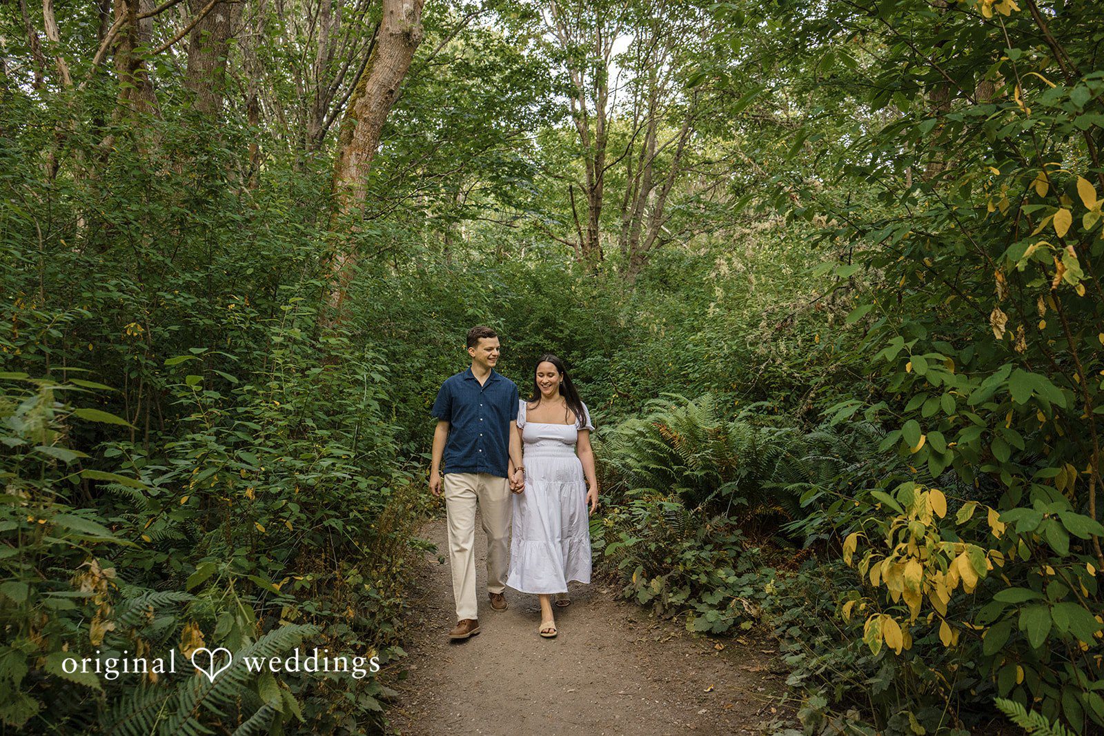 A couple standing together on a narrow dirt path enveloped by dense green trees.