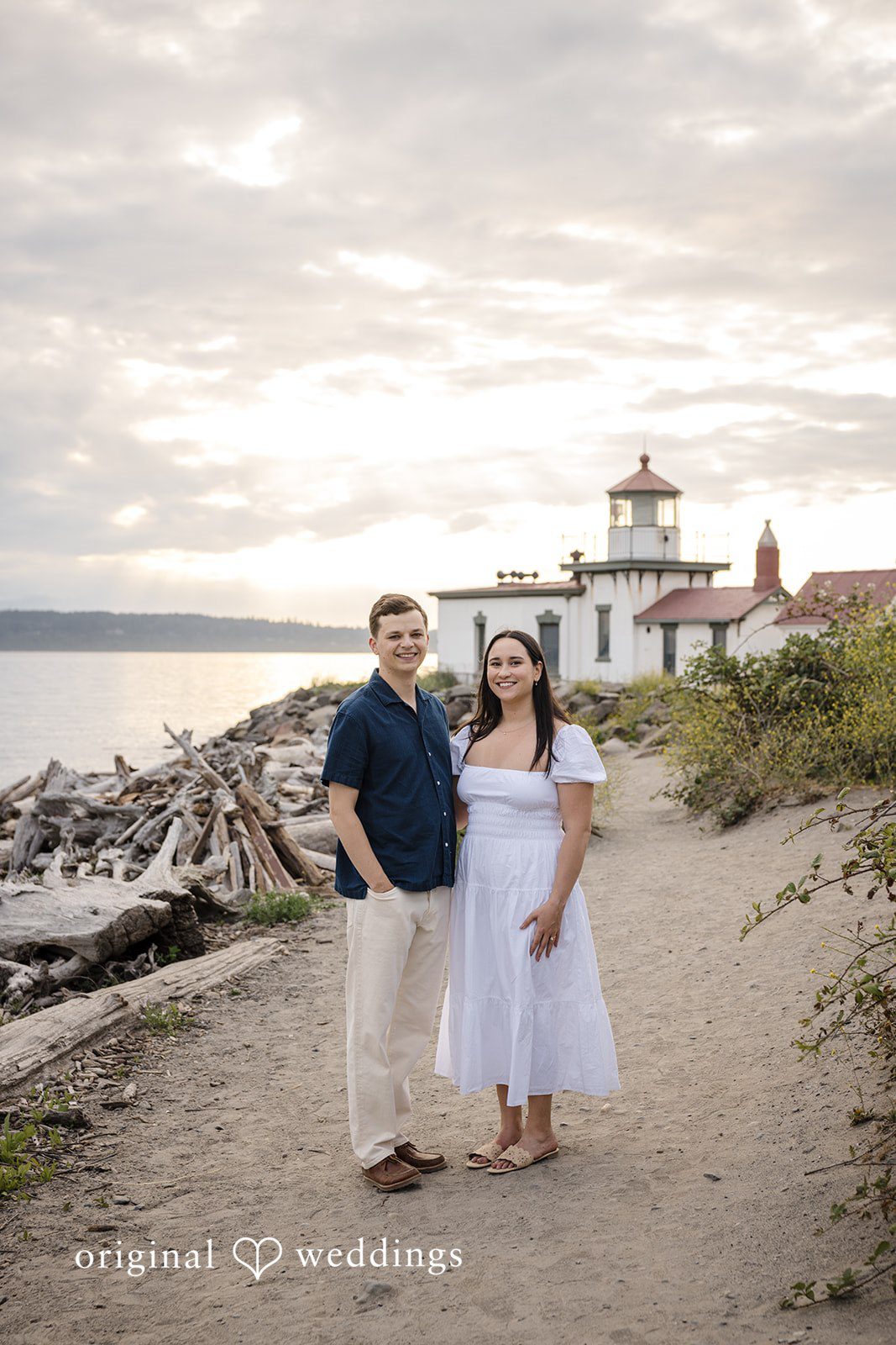 A couple standing together on a coastal trail with a lighthouse visible in the background.