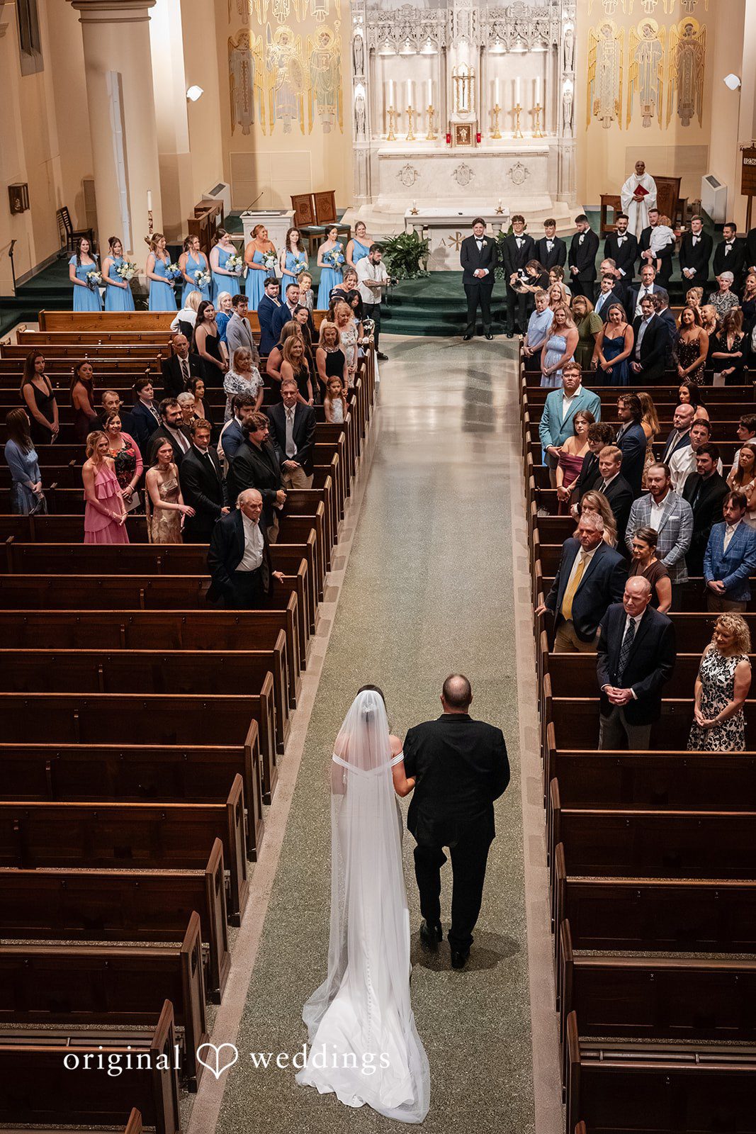 Mark + Caroline A wide view of a wedding procession walking down the church aisle filled with guests.