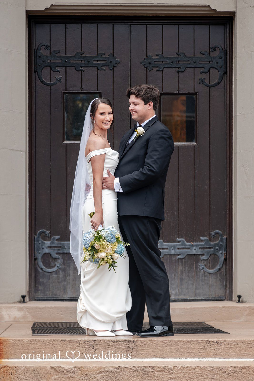 Mark + Caroline The bride and groom posing together in front of large wooden church doors.