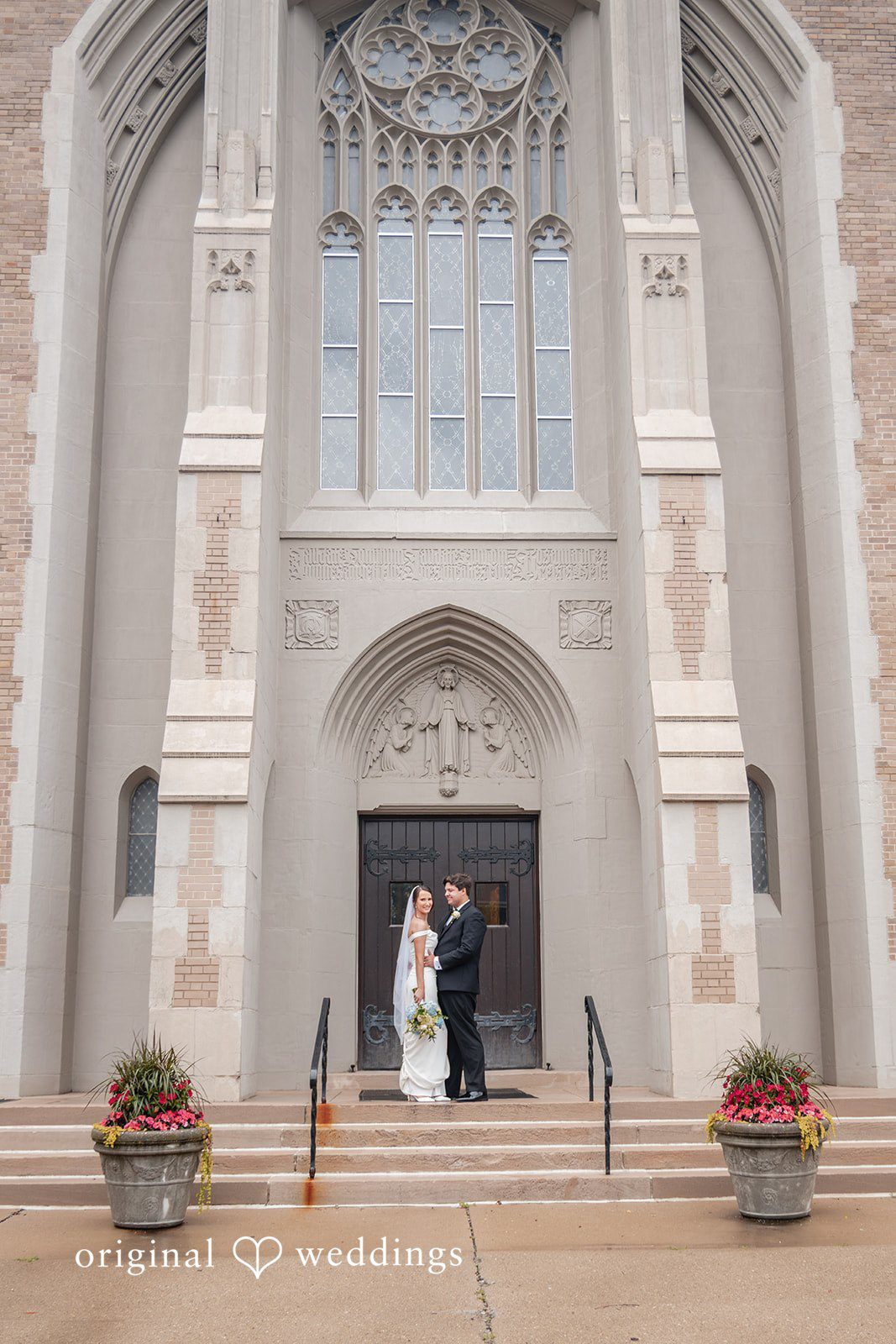 Mark + Caroline A wide shot of the bride and groom standing at the massive stone entrance of a church.