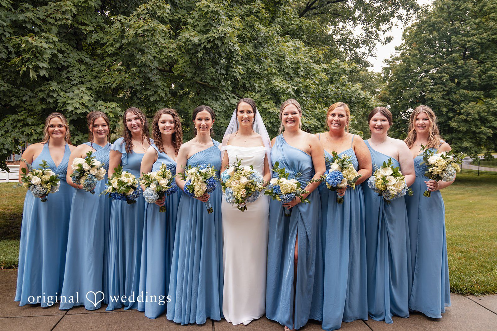 Mark + Caroline A bride posing with a large group of bridesmaids wearing dusty blue dresses.