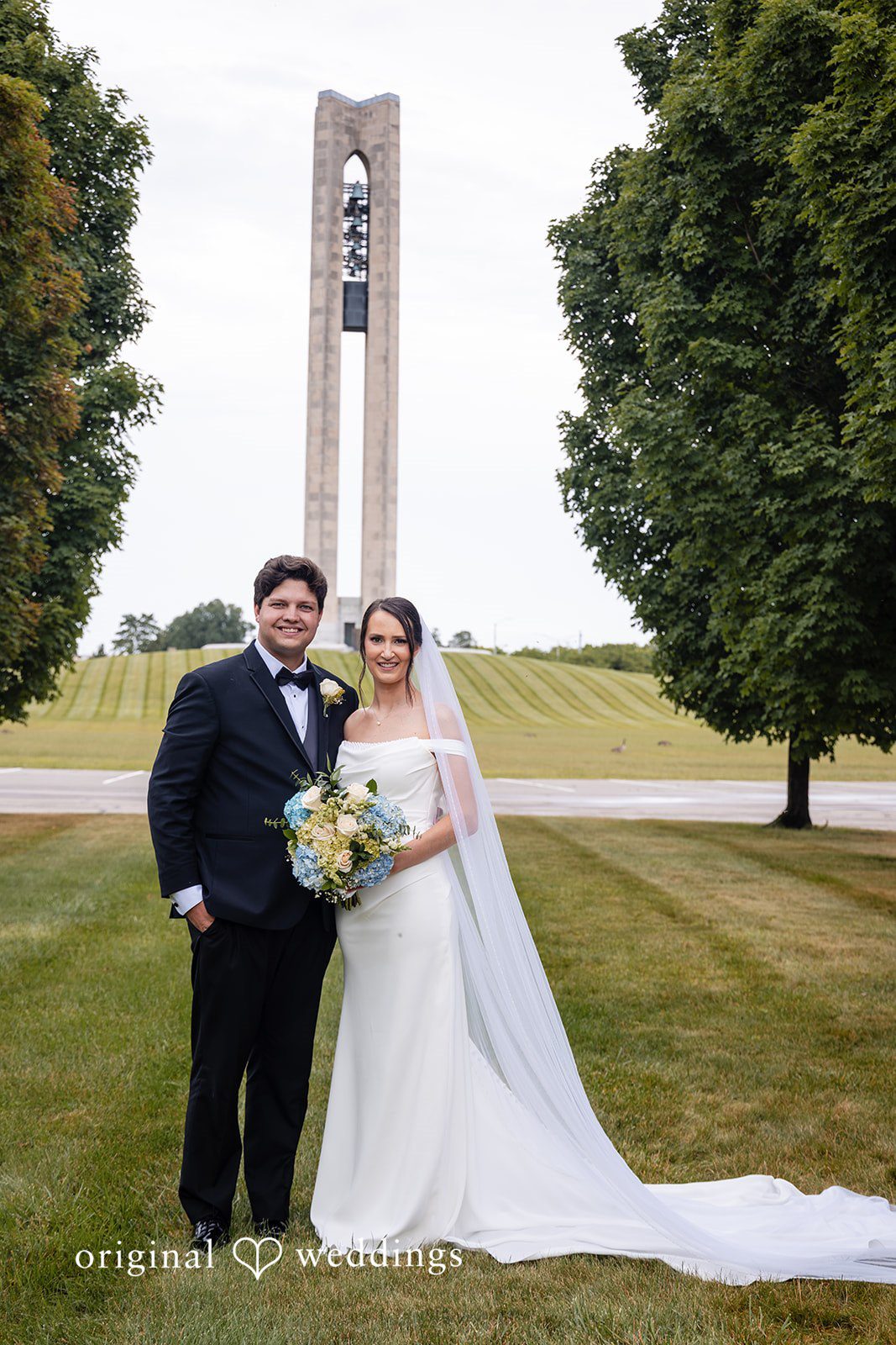 Mark + Caroline The bride and groom walking together across a grassy field with a monument in the background.
