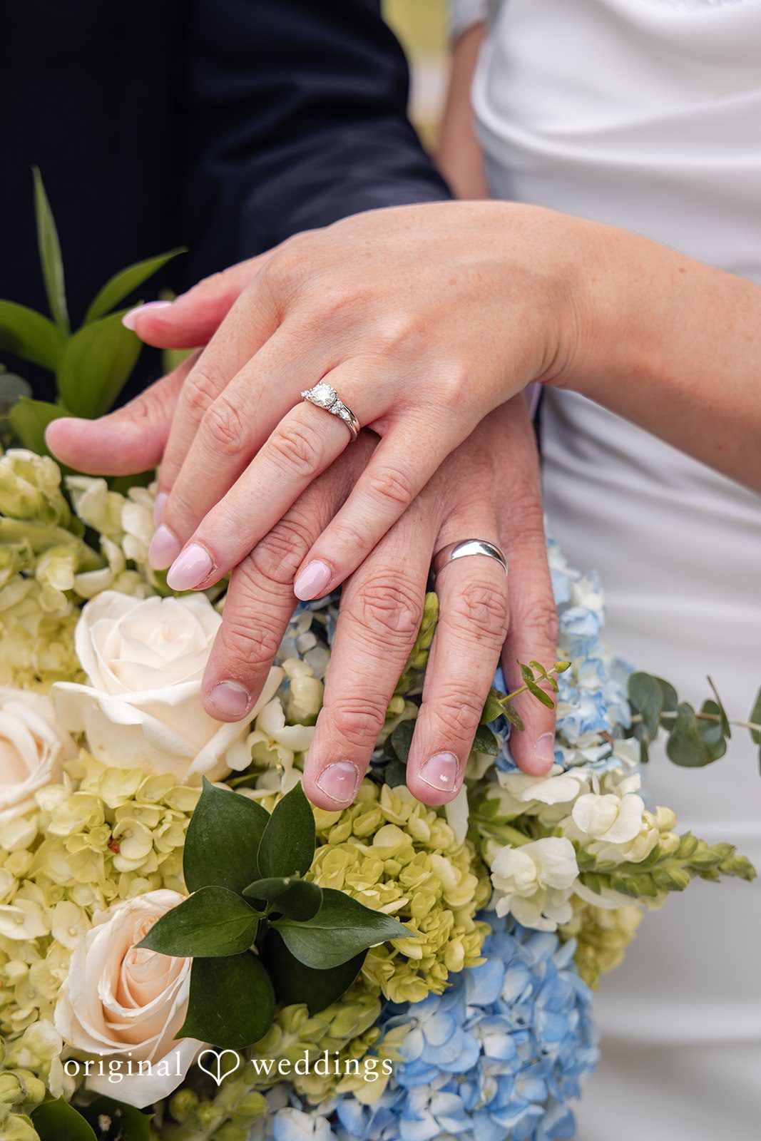 Mark + Caroline A close-up of the bride and groom's hands showcasing their wedding rings over a bouquet.