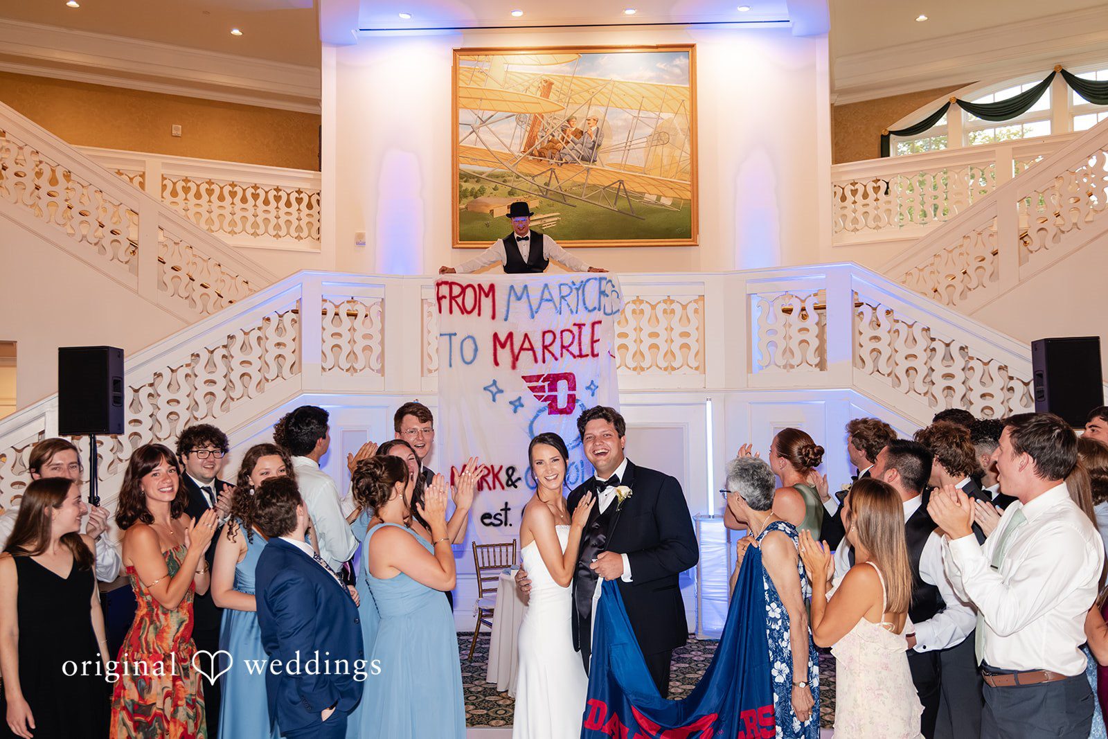 Mark + Caroline The newlyweds walking down a grand staircase into a reception hall filled with cheering guests.