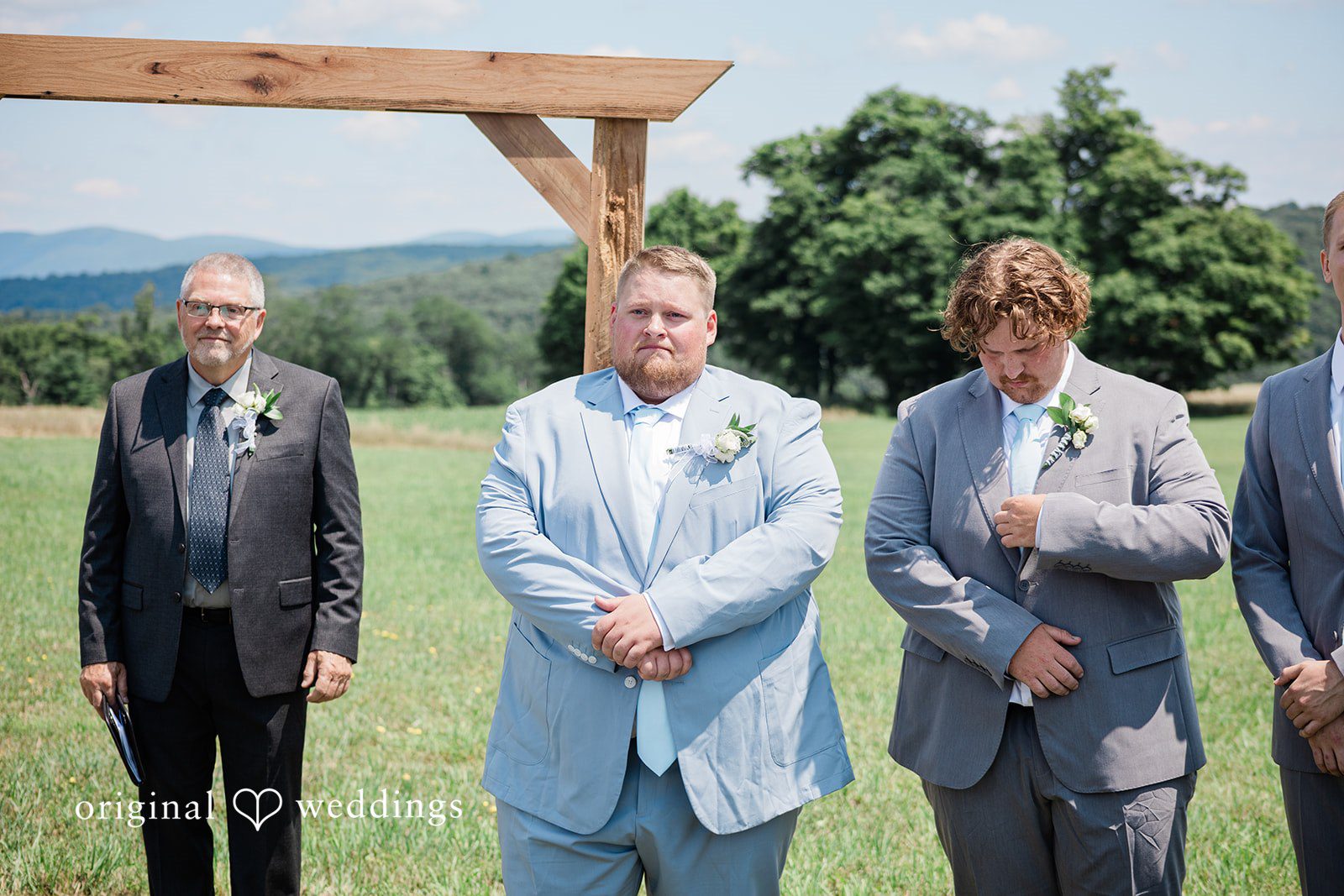 Michaela + Jake Three men in suits stand near a wooden arch during a wedding ceremony.