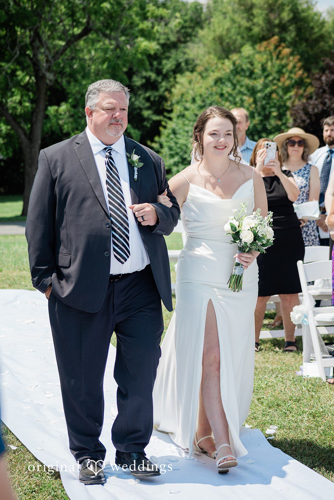 Michaela + Jake A man and a bride walk down a white aisle runner together.