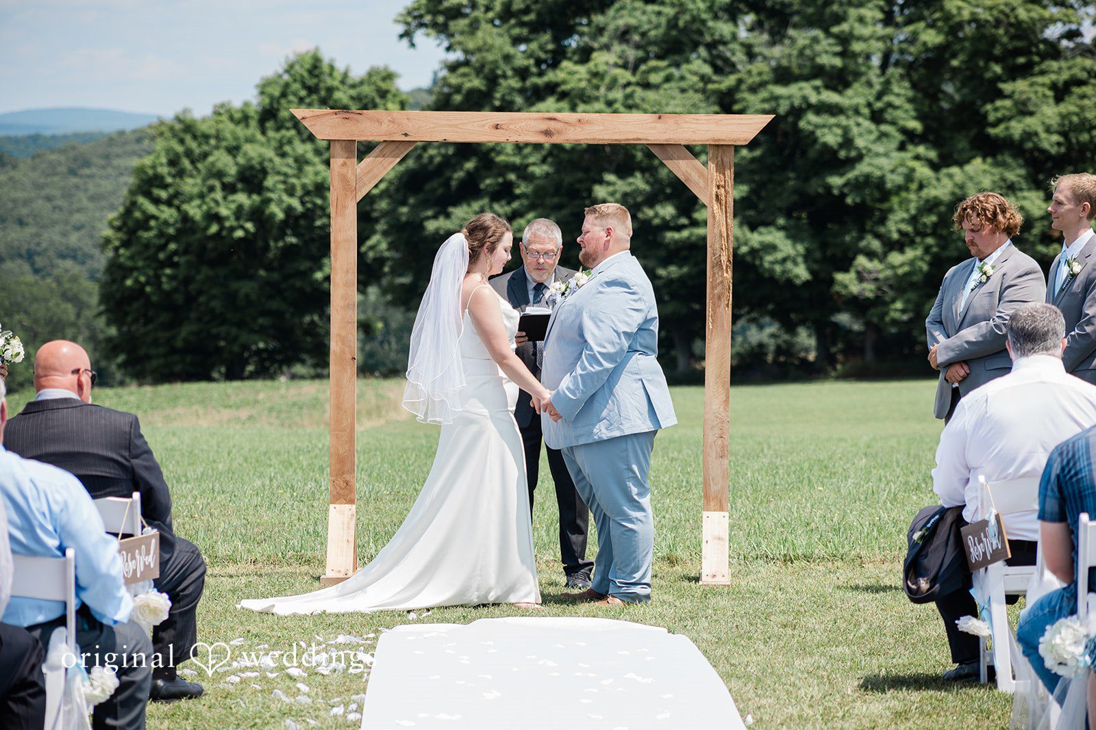 Michaela + Jake A wedding ceremony takes place under a wooden arch in an open field.