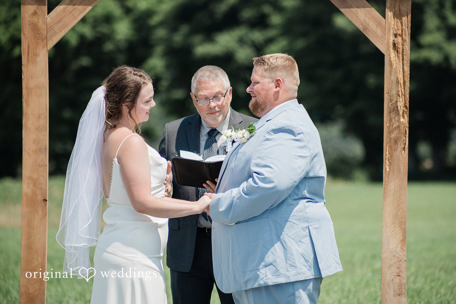 Michaela + Jake A bride and groom hold hands while facing an officiant during a ceremony.