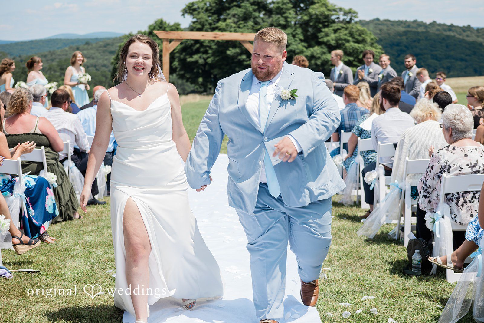 Michaela + Jake A newly married bride and groom walk hand-in-hand down the white aisle.