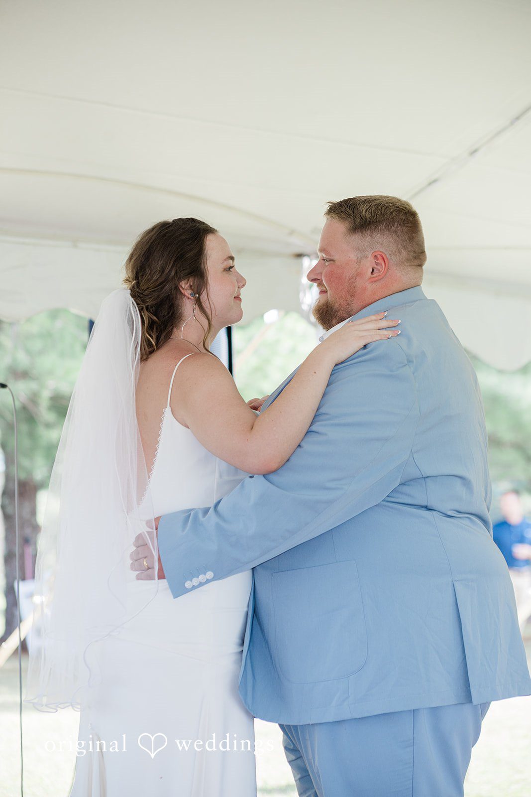 Michaela + Jake A bride and a groom in a light blue suit look lovingly at each other while embracing.