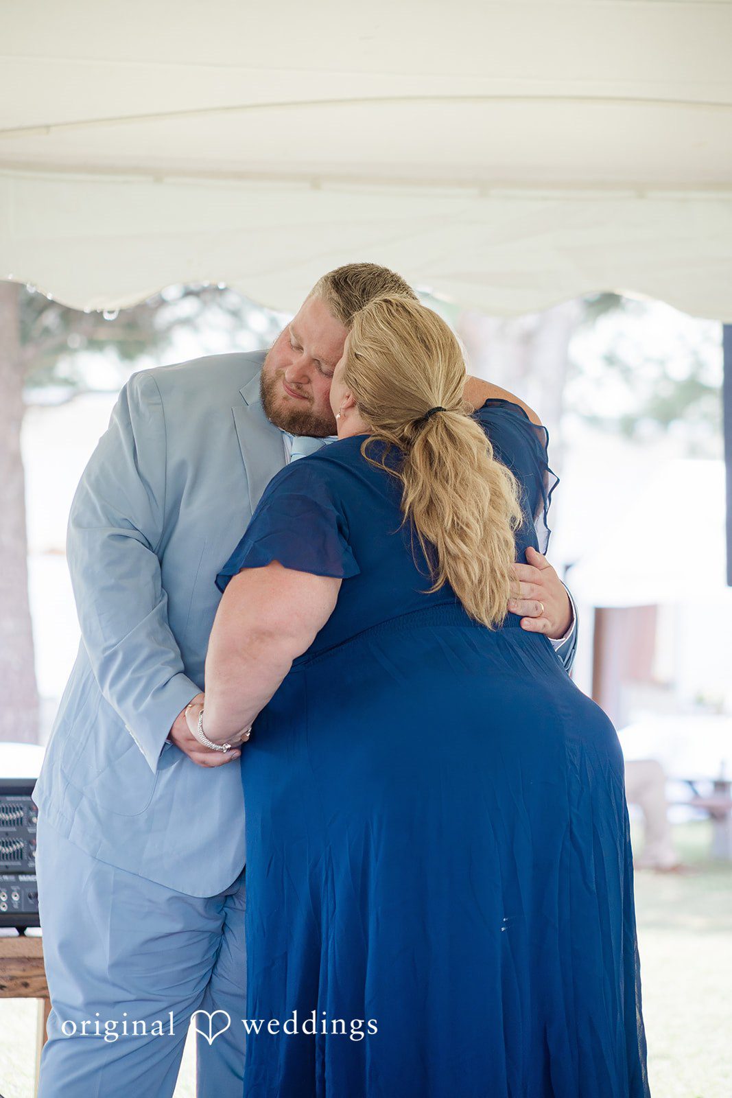 Michaela + Jake A groom in a light blue suit shares an emotional embrace with a woman in a dark blue dress.