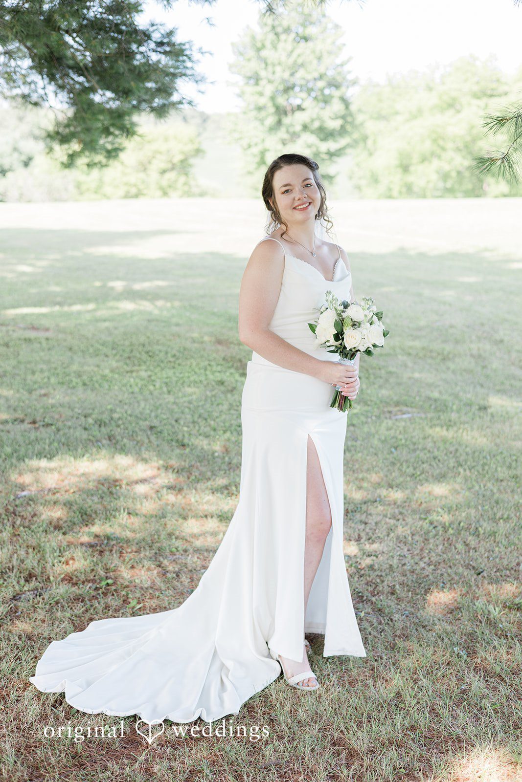 Michaela + Jake A bride in a high-slit white gown stands on grass holding a bouquet.