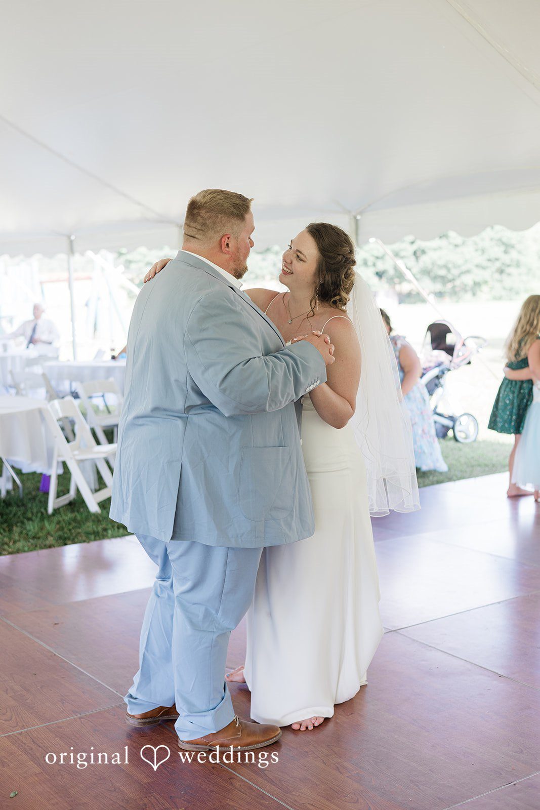 Michaela + Jake A groom in a light blue suit and a bride dance together on a wooden dance floor under a white tent.