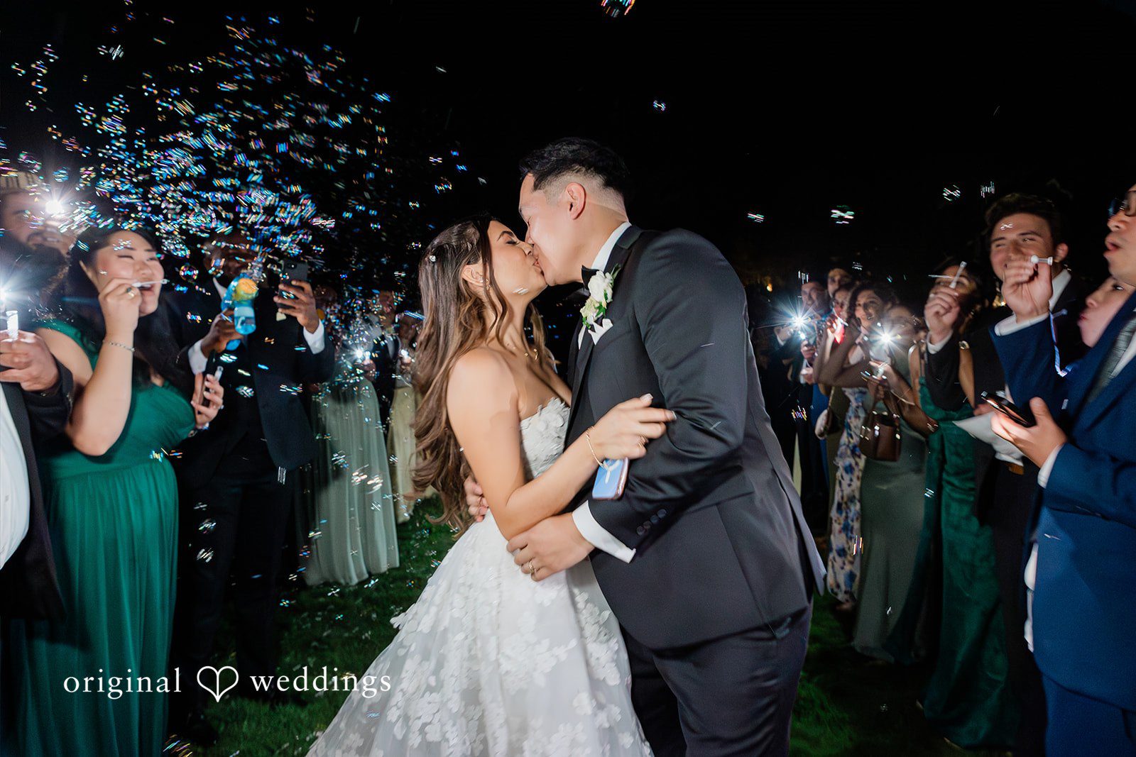 Bride and groom kissing on the dance floor surrounded by wedding guests.