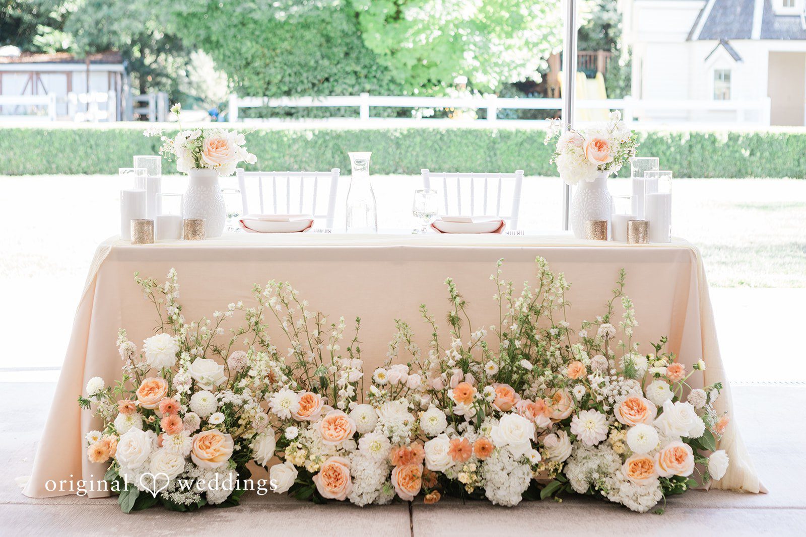 Reception head table decorated with light pink fabric and dense floral arrangements.
