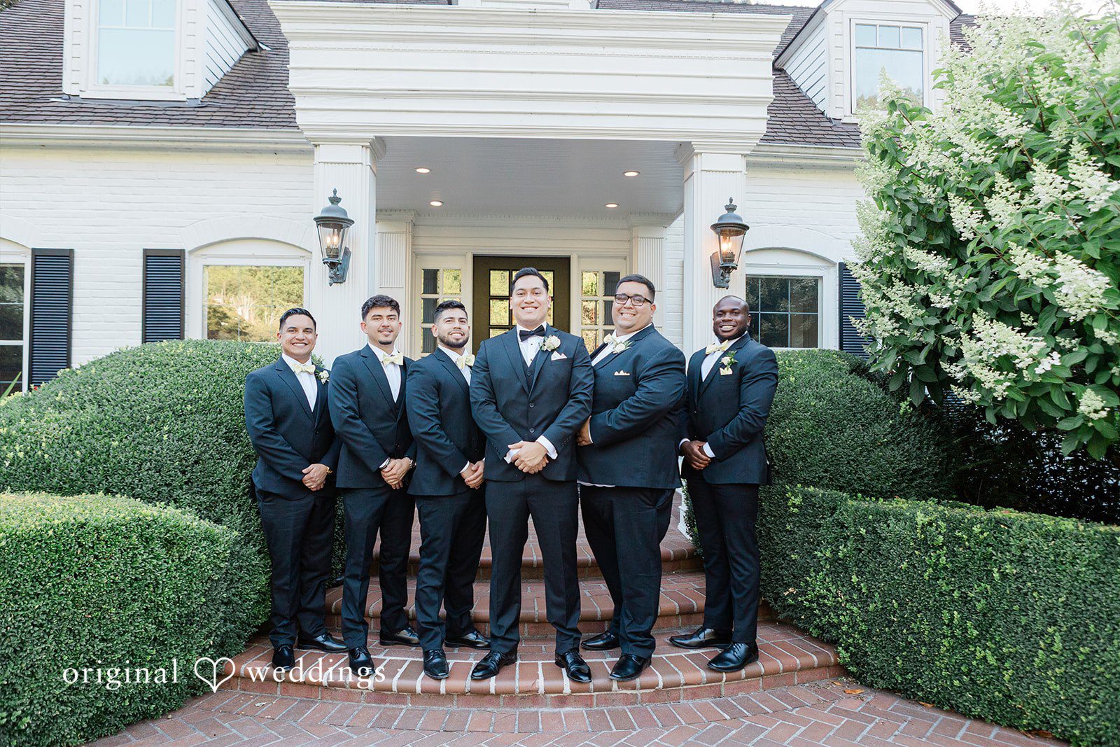 Group of groomsmen in dark suits standing together near hedges.