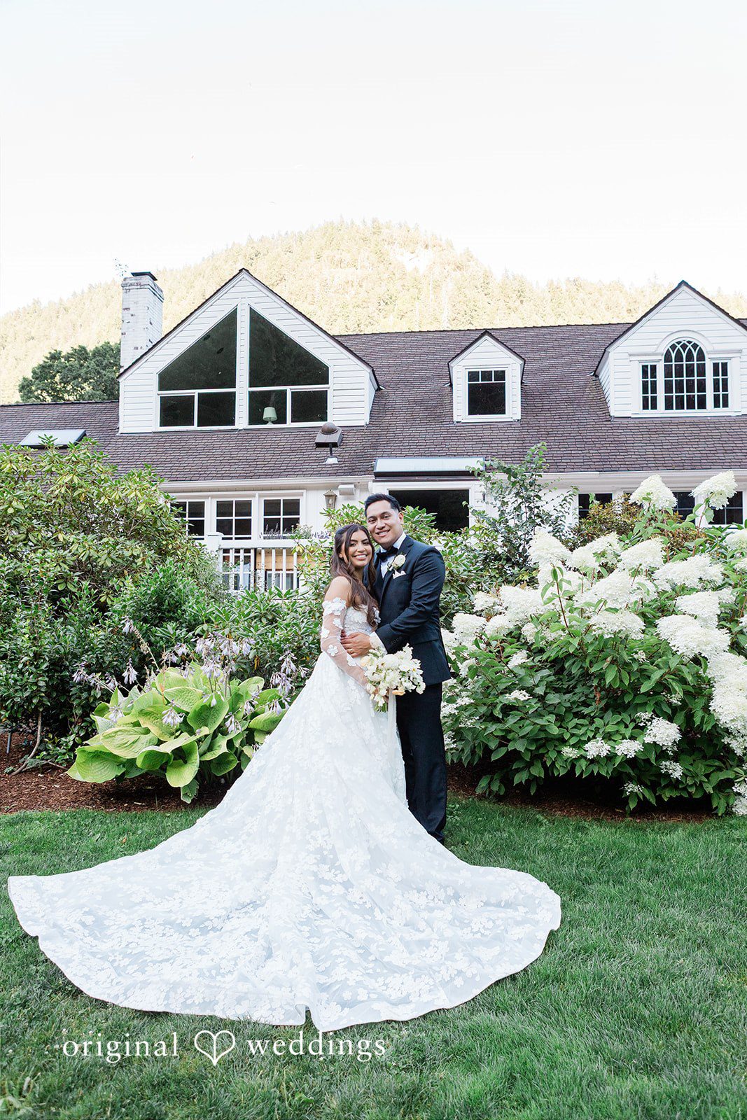 Bride and groom embracing on a lawn near a large house.
