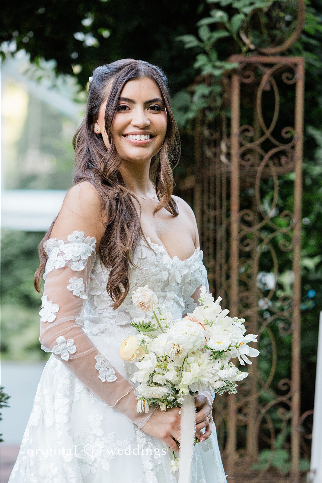 Smiling bride in a lace gown holding a white bouquet.