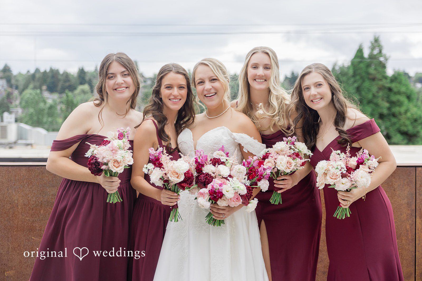 Michelle + Eric A bride smiling alongside four bridesmaids dressed in burgundy dresses holding bouquets.