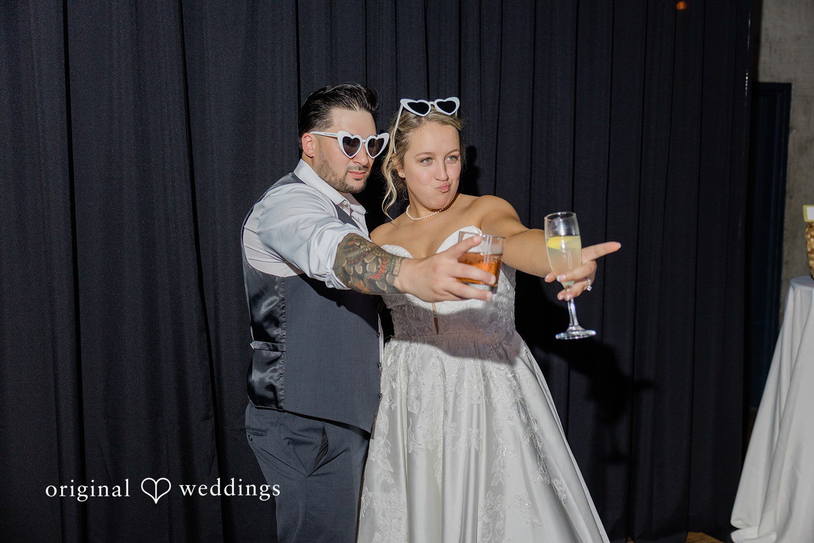 Michelle + Eric A bride and groom wearing sunglasses while holding up drinks against a dark backdrop.