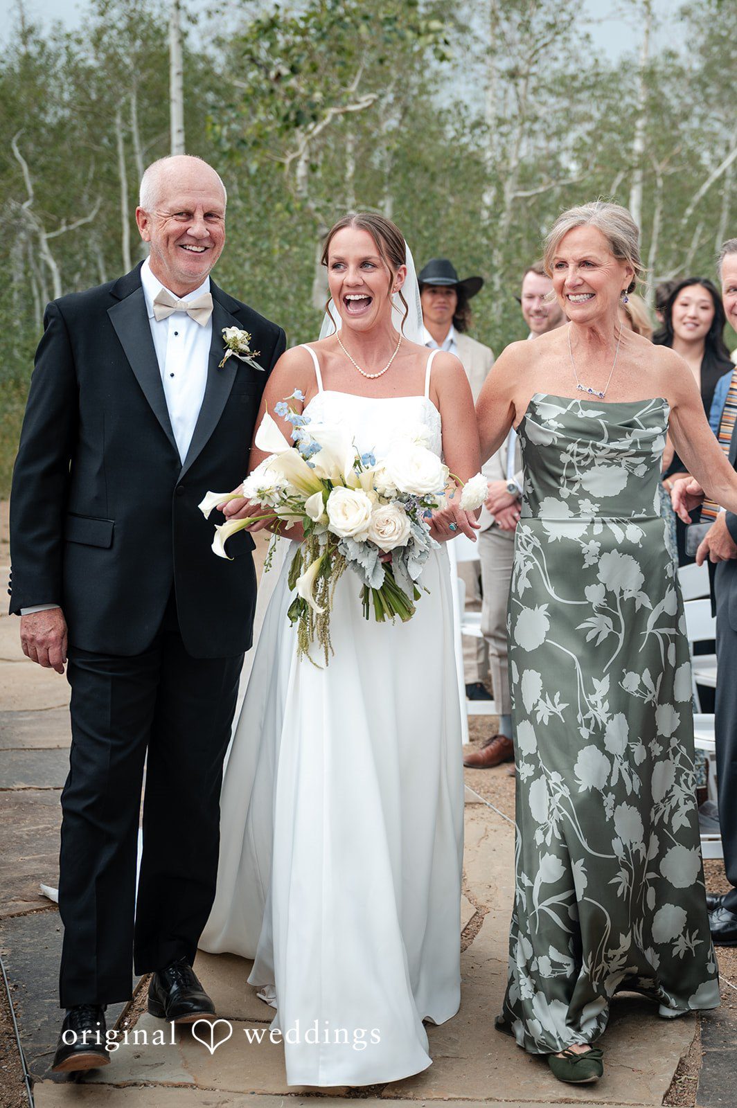 Emilia + Charlie A bride walking between two wedding guests holding a bouquet.