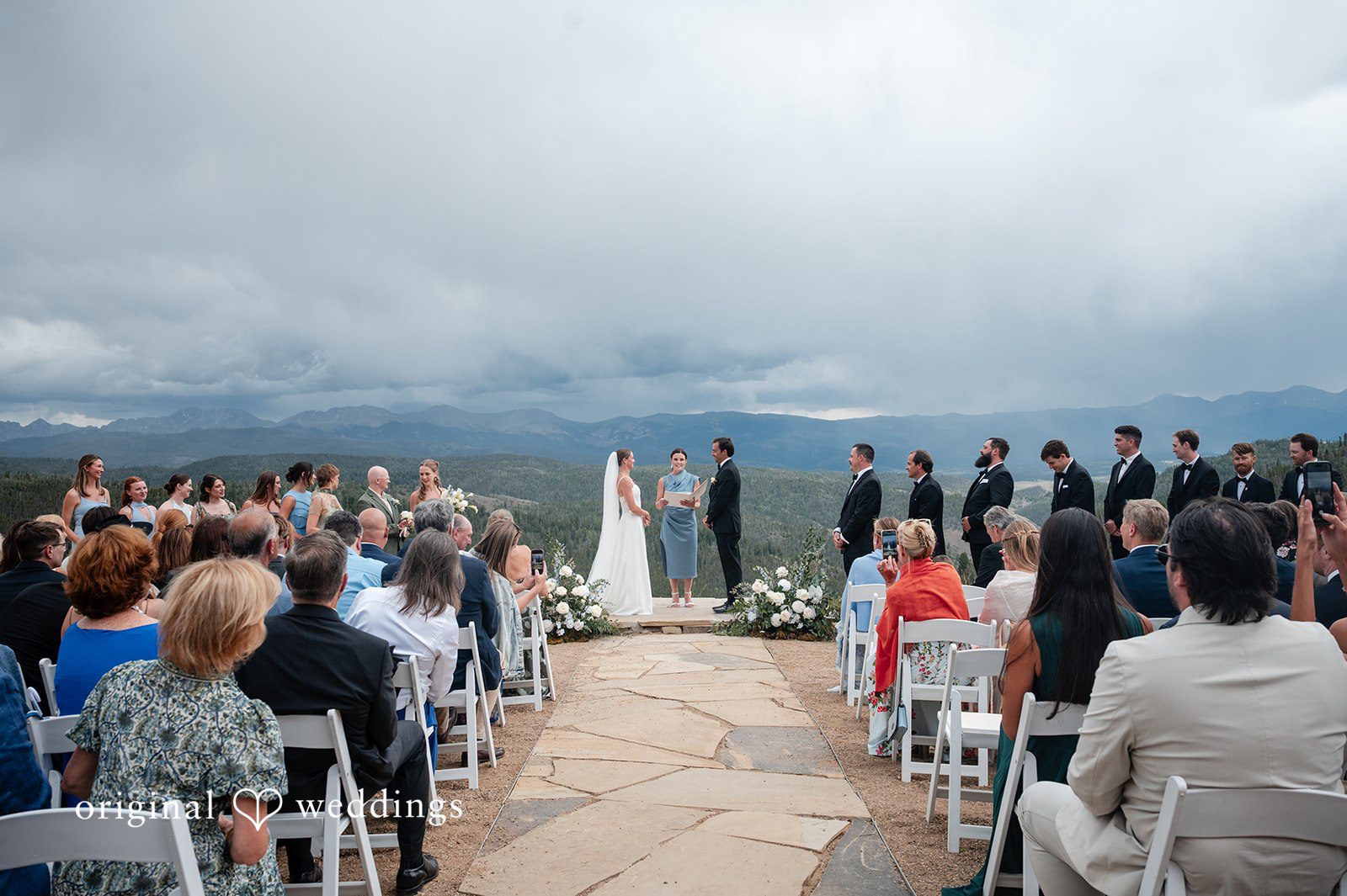 Emilia + Charlie An overview of guests seated at a wedding ceremony overlooking hills.