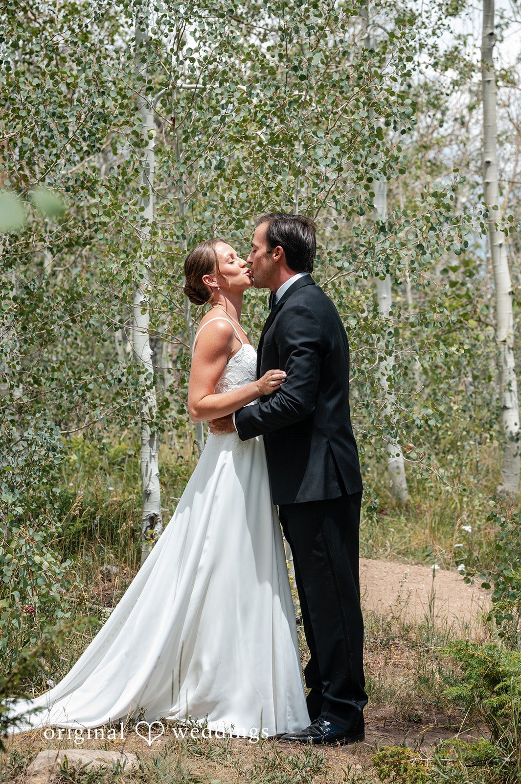 Emilia + Charlie A bride and groom sharing a kiss among tall trees.