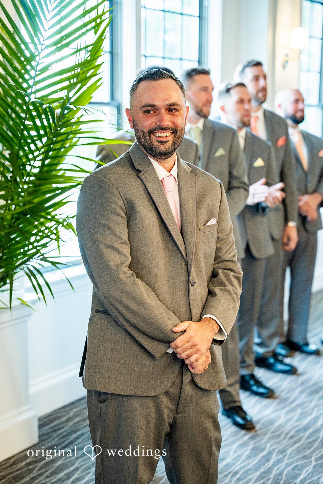 Alex + Courtney A groom smiling broadly in a grey suit next to green plants.