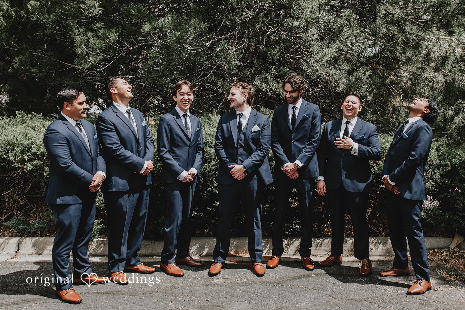 Groomsmen in matching dark suits posing together in front of a tree-lined backdrop.