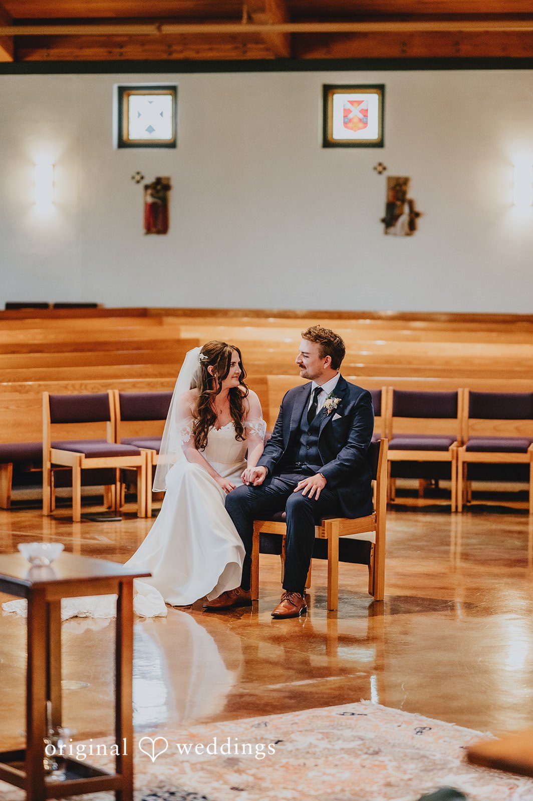 Bride and groom sitting together in wooden pews during their wedding ceremony.