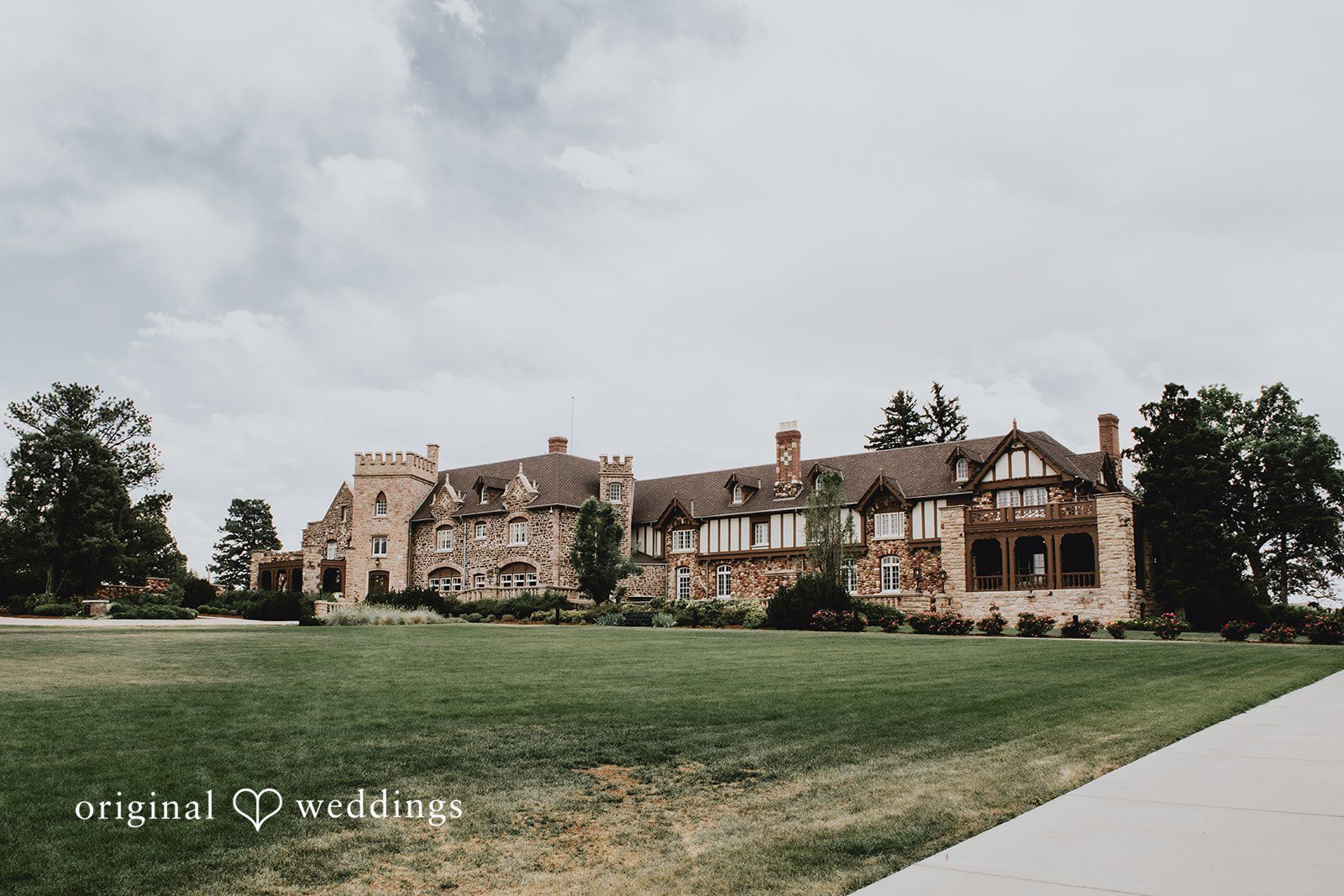 Large historic stone estate or manor with manicured green lawns under a cloudy sky.