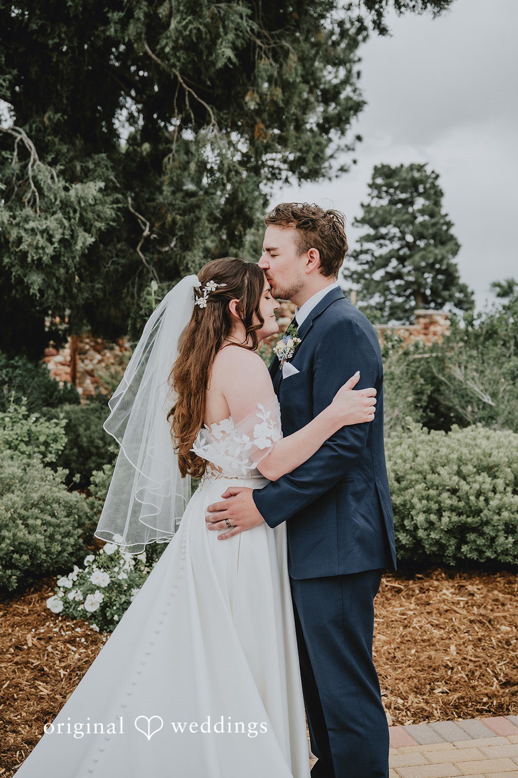 Groom kissing the bride from behind in front of a tree line.