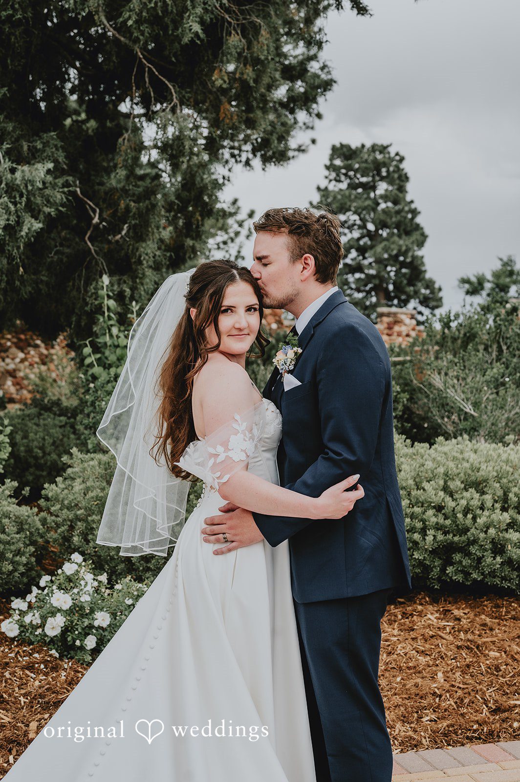 Bride and groom smiling and embracing against a background of evergreen trees.