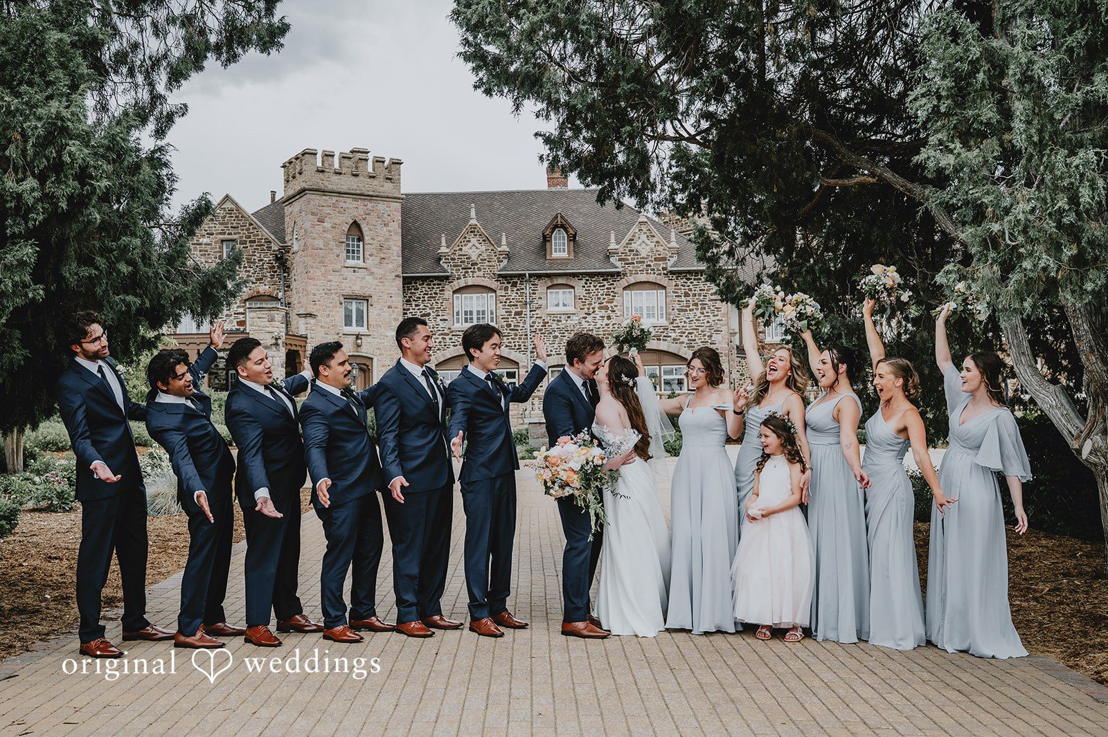 Large wedding party posing together in front of trees with a historic building behind them.